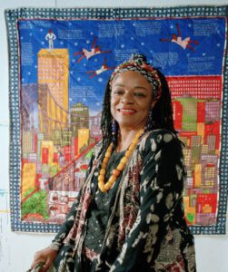image of Faith Ringgold standing before one of her multi-colored tapestries